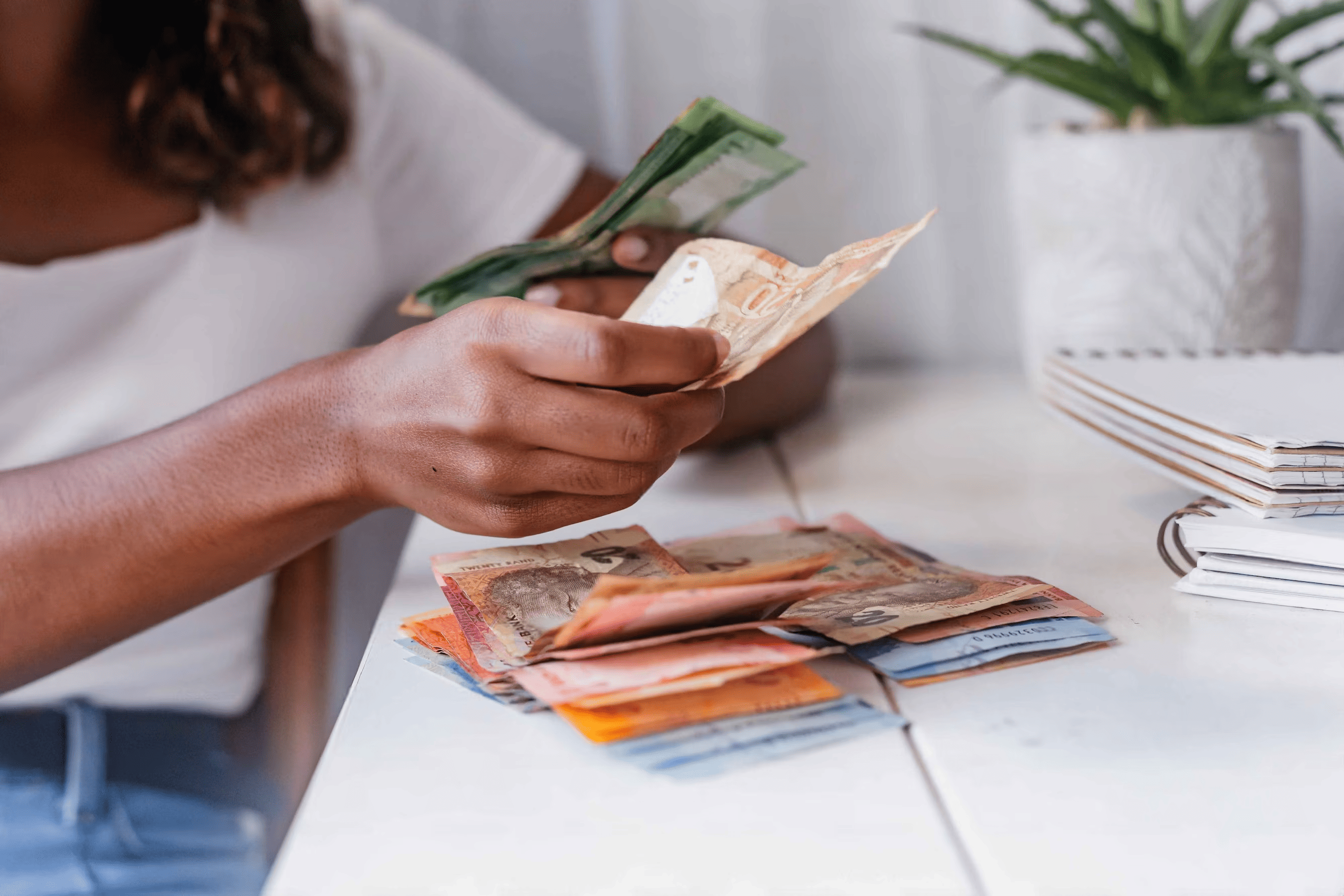 A woman sits at a table, counting South African Rands, with a stack of money spread out before her.