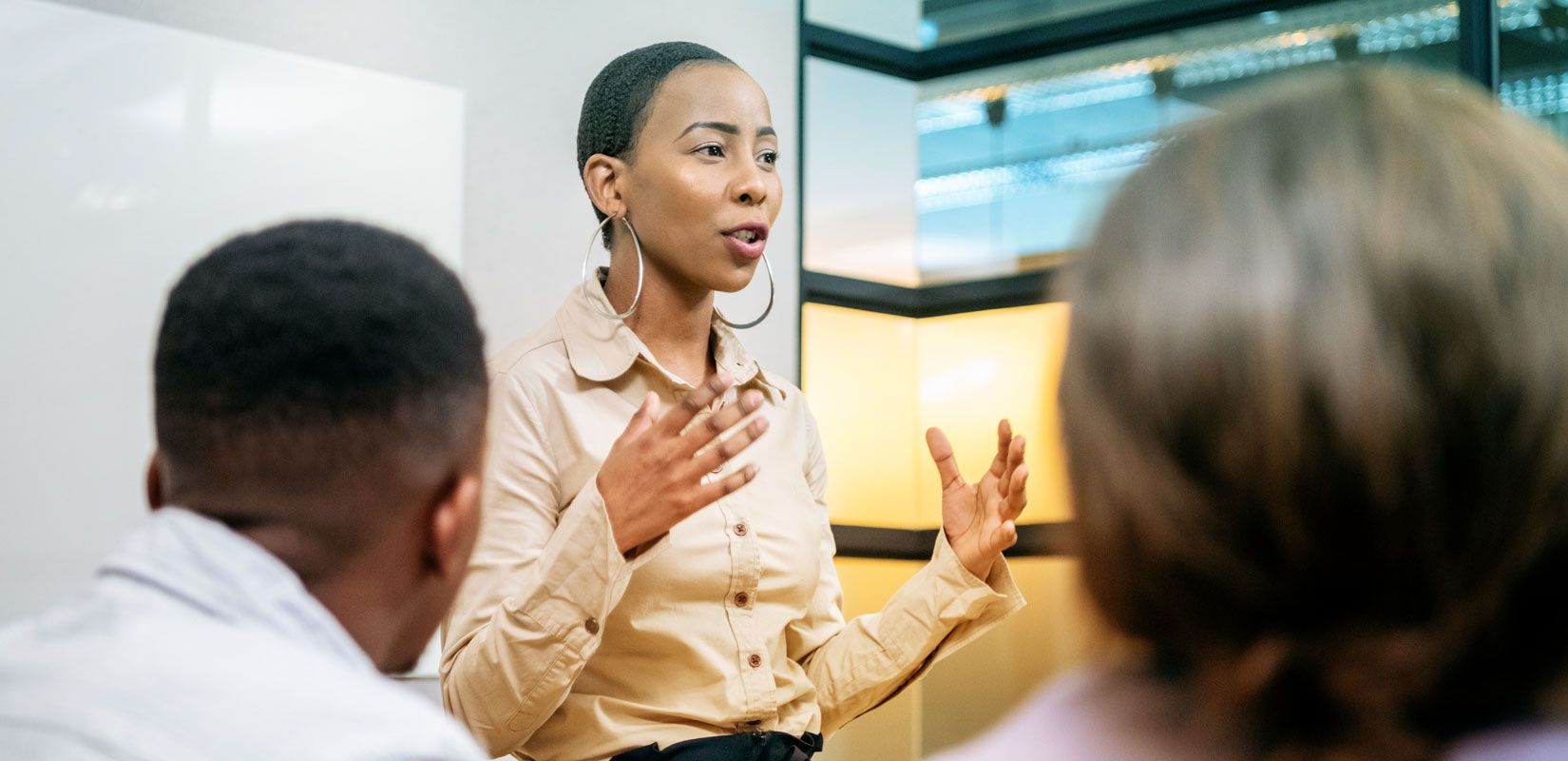 African lady standing and presenting in a boardroom, with two other people seated in the background, viewed from an over-the-shoulder perspective
