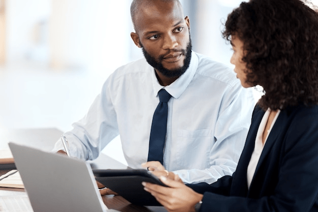 African businesswoman assisting an African businessman in formal wear beside a laptop, representing teamwork and professional support.